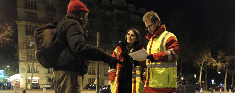 Photo de nuit montrant deux intervenants sociaux en gilets réfléchissants échangeant avec une personne dans la rue. La scène illustre une action de maraude.