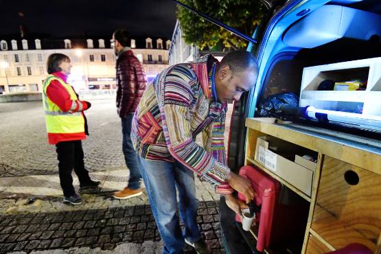 Photo de nuit montrant une action de maraude autour d’un véhicule aménagé. Une personne prépare une boisson chaude à l’arrière du véhicule, tandis que deux intervenants en gilet réfléchissant échangent à proximité.