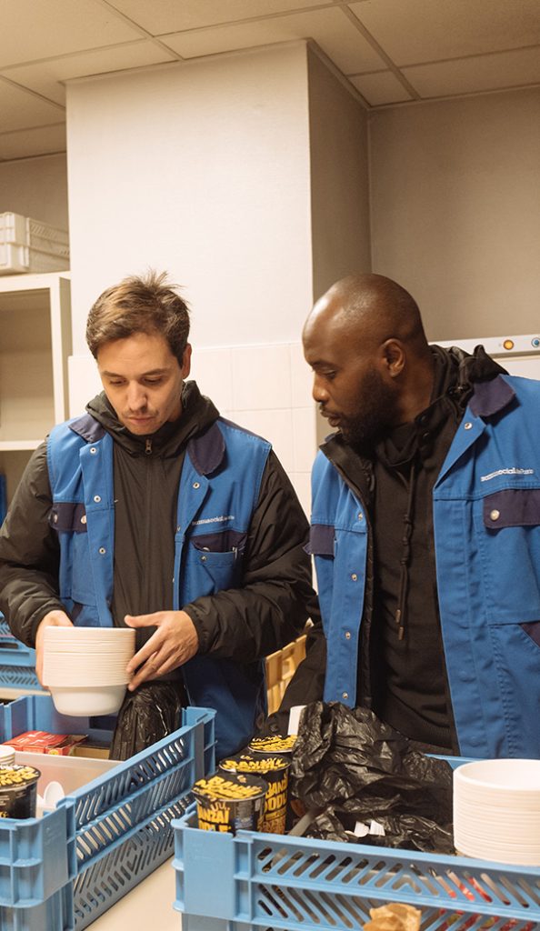 Photo montrant deux intervenants du Samu Social en gilets bleus préparant du matériel et des denrées alimentaires dans un local logistique. Des bols, des produits alimentaires et des caisses sont disposés devant eux.