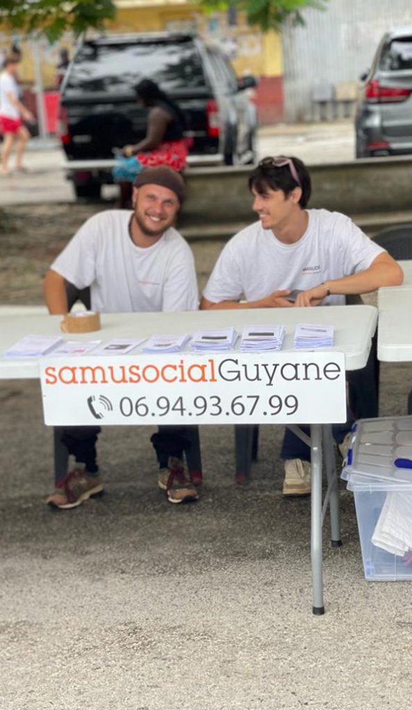 Photo montrant deux bénévoles assis derrière un stand du Samusocial Guyane, avec des documents d’information posés sur la table et une affiche indiquant le numéro de contact 06 94 93 67 99.