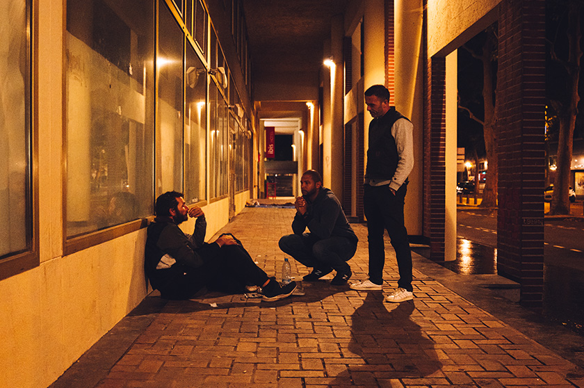 Photo prise dans une rue de nuit, avec trois hommes discutant. Un est assis contre un mur, une autre accroupi et l'autre debout les mains dans les poches.