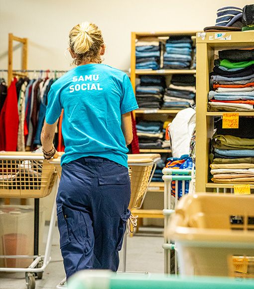 Photo montrant une personne portant un t-shirt “Samu Social” en train de circuler avec un chariot dans un espace de vestiaire solidaire. Des vêtements pliés et rangés sur des étagères entourent la scène.