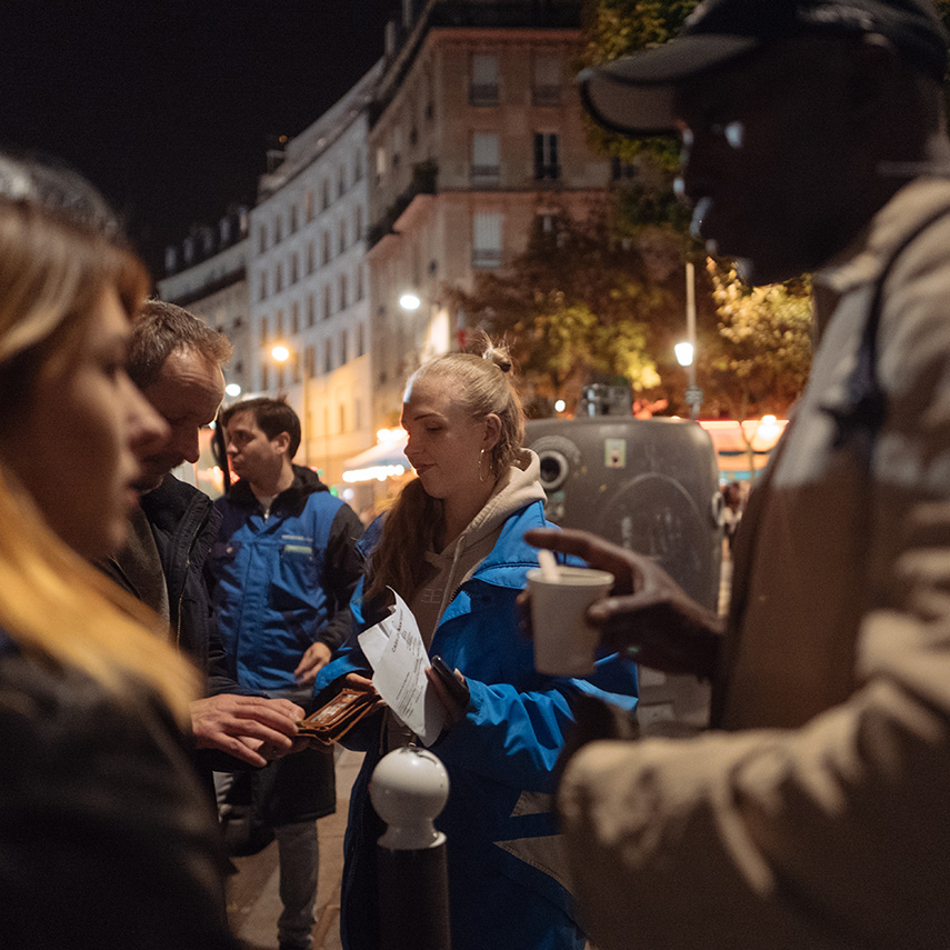 Photo de nuit montrant des intervenants du Samu social échangeant avec plusieurs personnes dans la rue. Une boisson chaude et des documents sont visibles.