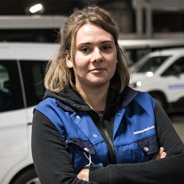 Portrait d’une intervenante du Samu Social portant un gilet bleu, les bras croisés, devant un véhicule de maraude.